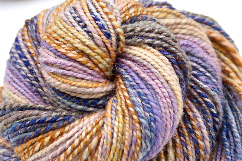 A close up view of a one of a kind, hand dyed Gradient skein of multicolored Orange, Gold, Light Peach, Lavender, Navy Blue, Gray, and Brown self-striping wool Yarn. 