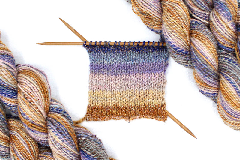 A sample swatch knitted from a one of a kind, hand dyed Gradient skein of multicolored Orange, Gold, Light Peach, Lavender, Navy Blue, Gray, and Brown self-striping wool Yarn. 