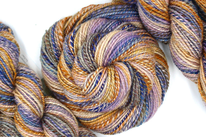A one of a kind, hand dyed Gradient skein of multicolored Orange, Gold, Light Peach, Lavender, Navy Blue, Gray, and Brown self-striping wool Yarn coiled attractively in the center of the frame. 