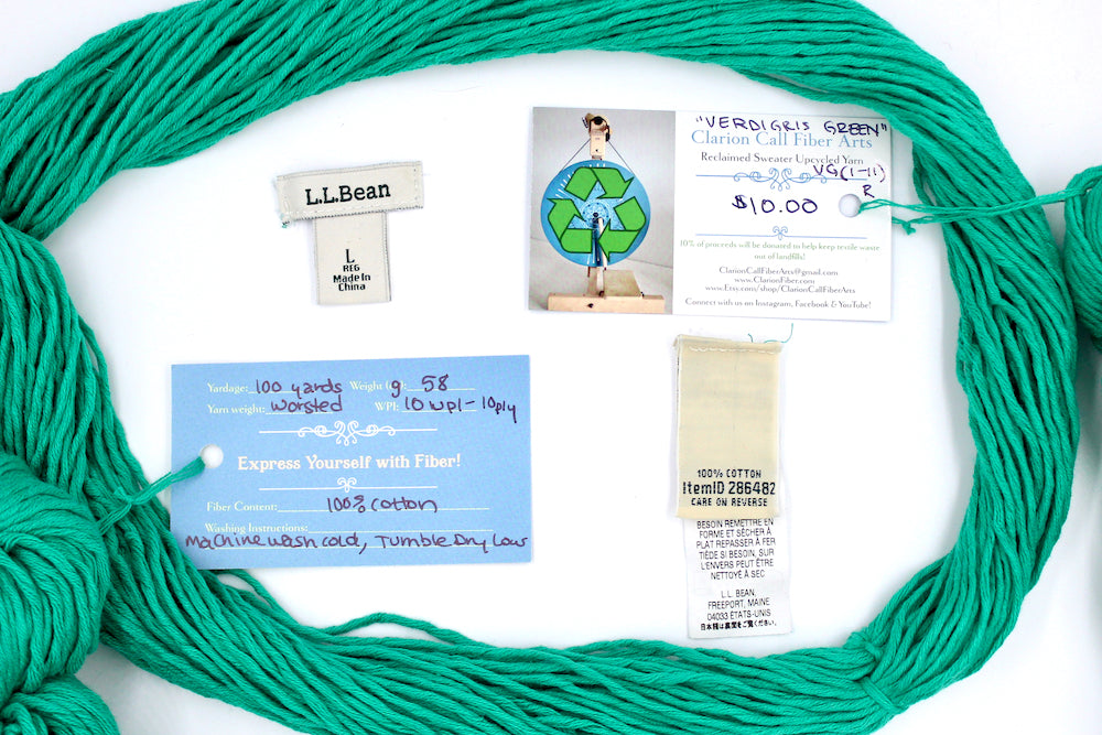 A skein of Vegan, Bright Blue Green, 100% Cotton, Worsted Weight yarn encircling the sweater labels it was recycled from, as well as the hand written yarn tags detailing the specs of this recycled yarn to help you make your next project! 