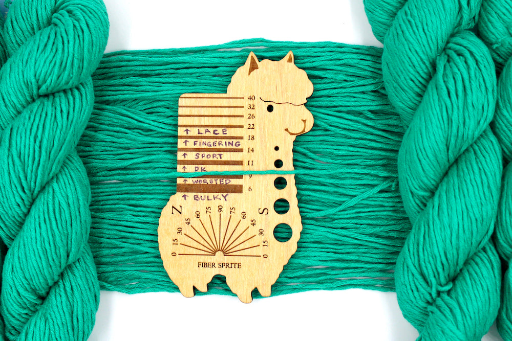 A skein of Vegan, Bright Blue Green, 100% Cotton, Worsted Weight Yarn recycled by hand from unwanted sweaters draped over the center of the frame with one strand on top of a Llama shaped Yarn weight tool, to better illustrate the thickness of the yarn. 