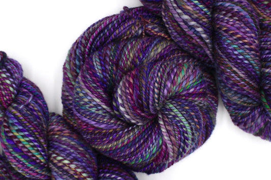 A one of a kind, Worsted Weight, hand dyed variegated skein of multicolored Purple, Fuchsia, Green, Navy Blue, Teal, and Pink self-striping wool Yarn coiled attractively in the center of the frame.