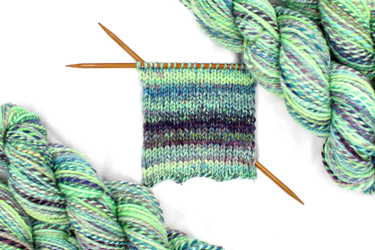 A sample swatch knitted on size 5 knitting needles from a one of a kind, Worsted Weight, hand dyed variegated skein of multicolored Lime Green, Yellow, Seafoam, Navy, Blue, and Purple self-striping 100% Superwash Merino wool Yarn. 