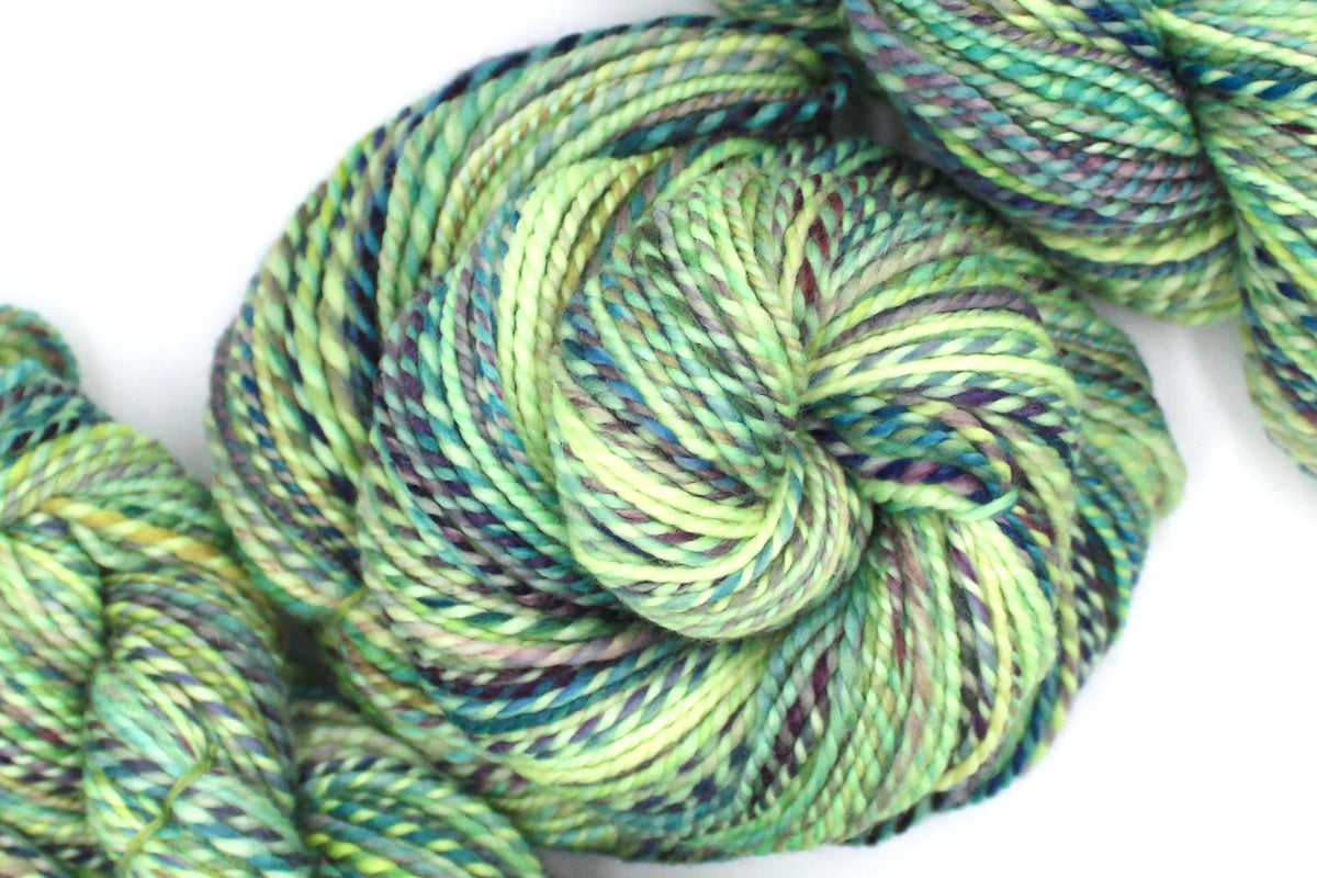 A one of a kind, Worsted Weight, hand dyed variegated skein of multicolored Lime Green, Yellow, Seafoam, Navy, Blue, and Purple self-striping 100% Superwash Merino wool Yarn coiled attractively in the center of the frame. 
