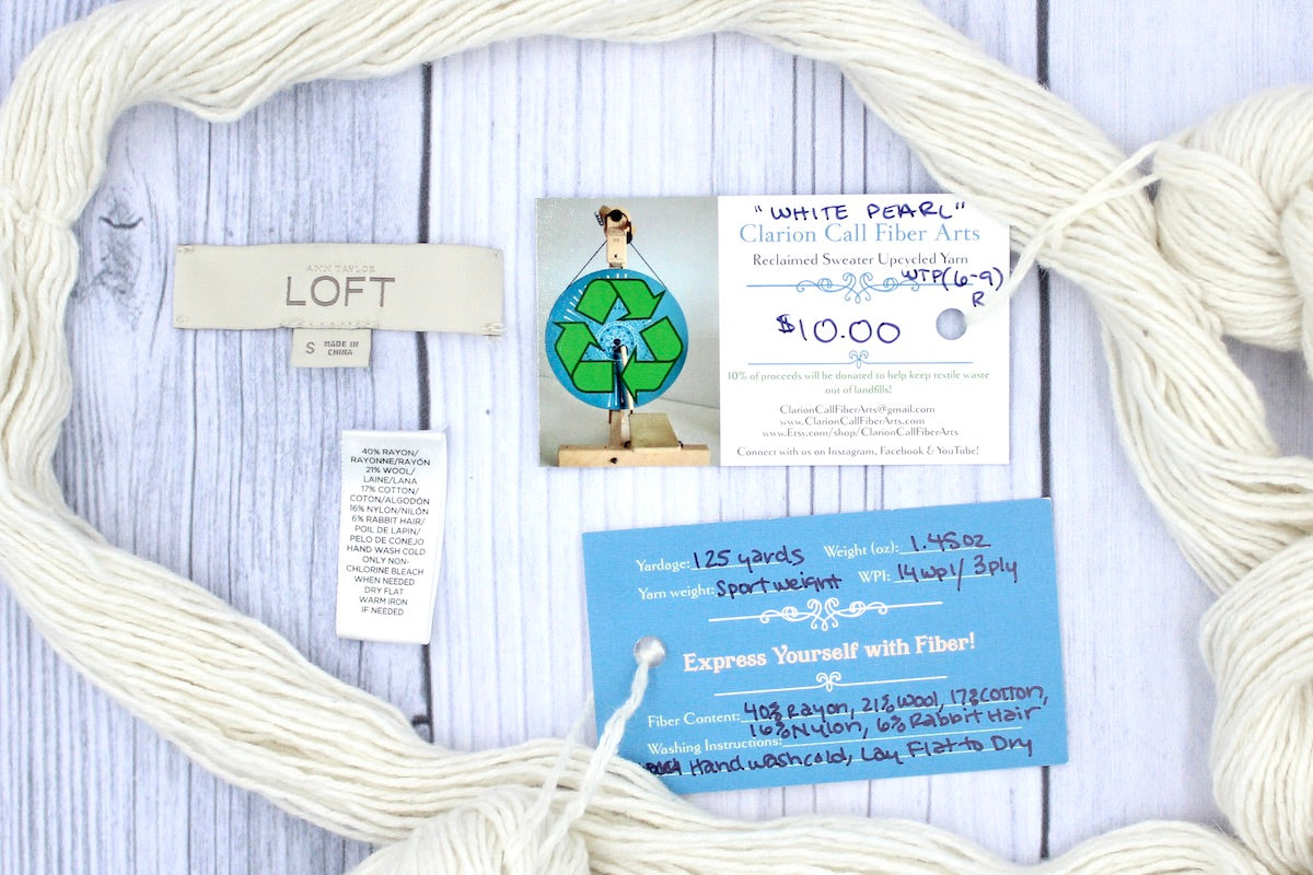 A skein of Cream/ Off White/ Ecru/ Pearl, Rayon/ Wool/ Cotton/ Nylon/ Rabbit Hair, Sport Weight yarn encircling the sweater labels it was recycled from, as well as the hand written yarn tags detailing the specs of this upcycled yarn to help you make your next project! 