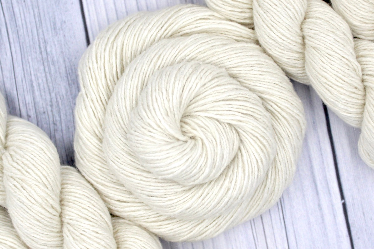 A skein of Cream/ Off White/ Ecru/ Pearl, Rayon/ Wool/ Cotton/ Nylon/ Rabbit Hair, Sport weight Yarn recycled by hand from unwanted sweaters swirled attractively in the center of the frame. 