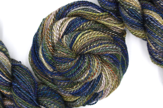 A one of a kind, hand dyed Gradient skein of multicolored Navy Blue, Indigo, Dark Green, Brown, Gold, and Taupe self-striping wool Yarn coiled attractively in the center of the frame. 