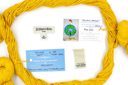 A skein of Vegan, Mustard Yellow, 100% Cotton, Sport Weight yarn encircling the sweater labels it was recycled from, as well as the hand written yarn tags detailing the specs of this recycled yarn to help you make your next project! 
