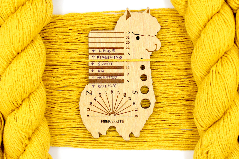 A skein of Vegan, Mustard Yellow, 100% Cotton, Sport Weight Yarn recycled by hand from unwanted sweaters draped over the center of the frame with one strand on top of a Llama shaped Yarn weight tool, to better illustrate the thickness of the yarn. 